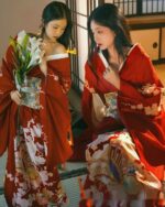 Two women wearing a vibrant red aesthetic kimono with floral patterns. They hold lilies and appear to be indoors.