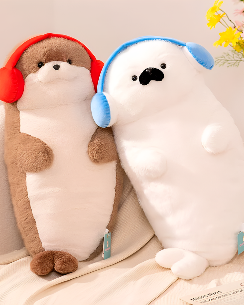 Two plush toys: a brown Kawaii Seal Plushie and a white otter with headphones, displayed on a textured blanket.