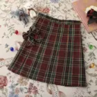 A red and green JK Plaid Skirt with a bow detail, displayed on a floral background.