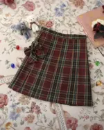 A red and green JK Plaid Skirt with a bow detail, displayed on a floral background.