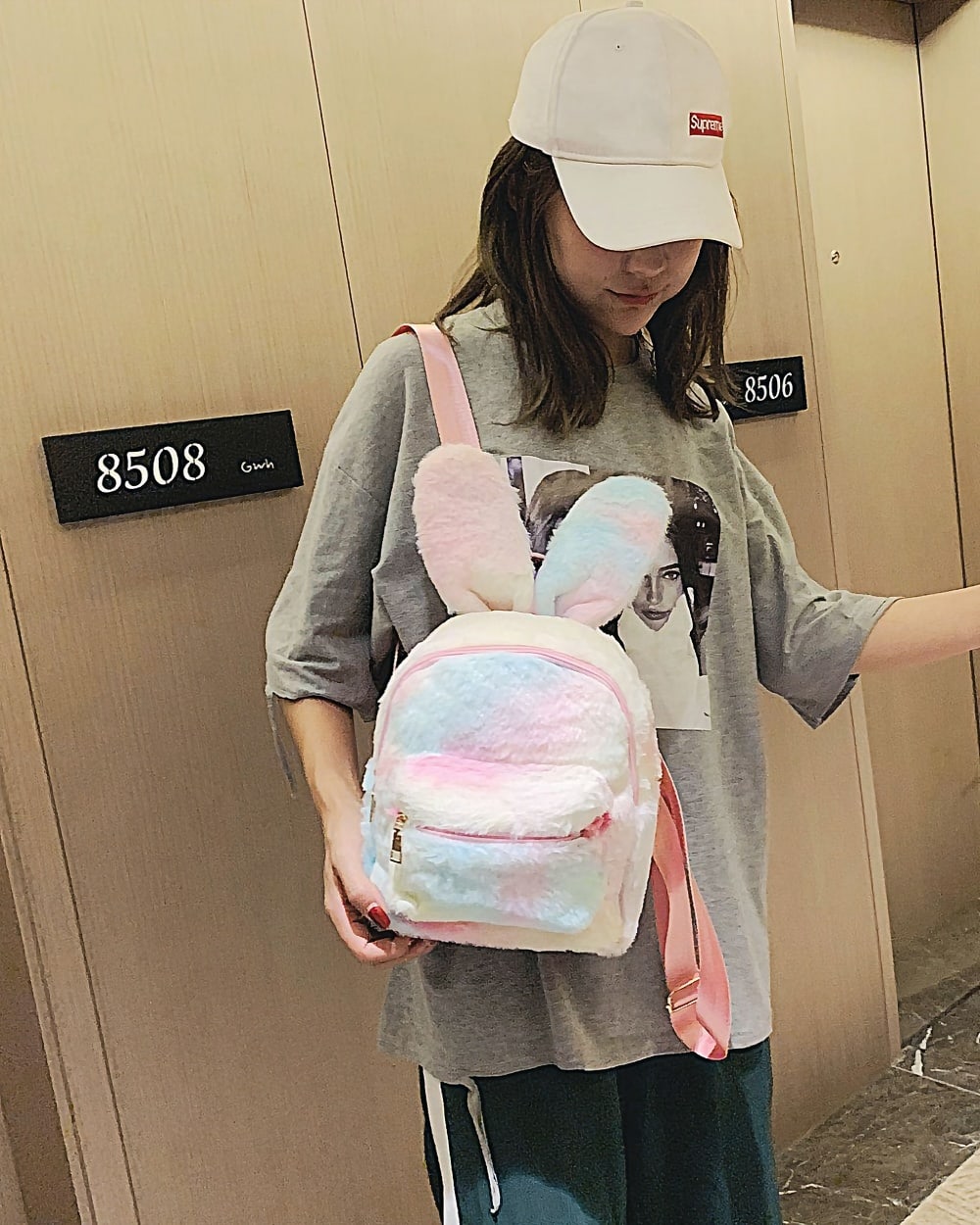 A pink and white Rabbit Ears Plush Backpack is worn by a person in a public space.