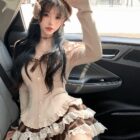 Model wearing a coquette aesthetic outfit: ruffled top and plaid skirt with lace details, alongside a hat.