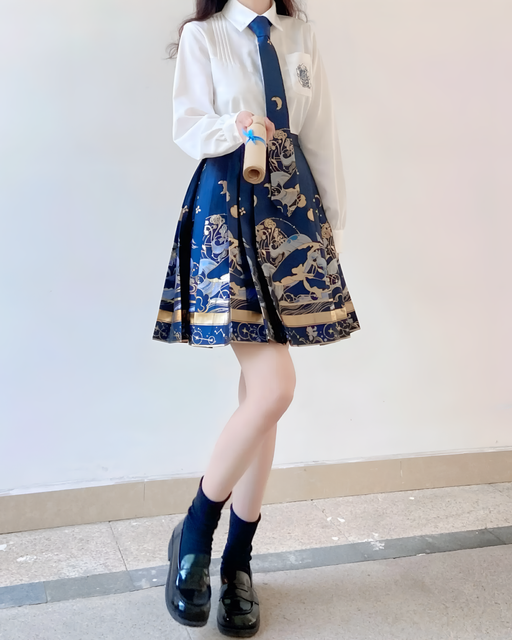 Woman wearing a white blouse and navy blue celestial pleated skirt with a patterned design. Dark socks and black shoes.