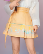 Model wearing a yellow and blue celestial pleated skirt with floral print and tie waist. A detailed view of the celestial pleated skirt.