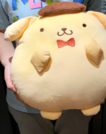 Hands holding a large, round Chonky Purin Plushie with a sweet expression and a red bow tie. Soft and huggable!