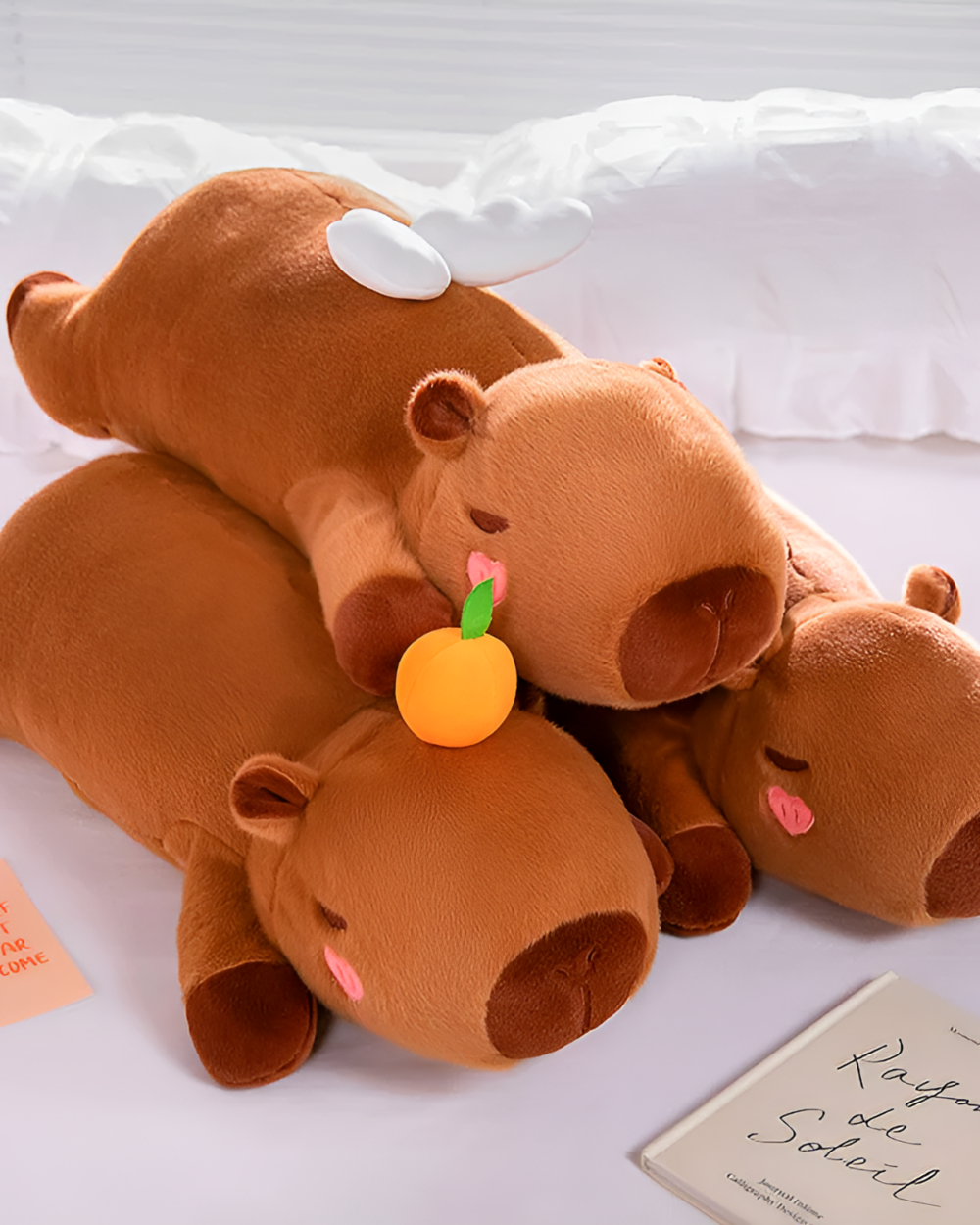 Chonky brown capybara plushie resting on a bed with a small orange. A cozy and adorable capybara plushie.