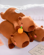 Chonky brown capybara plushie resting on a bed with a small orange. A cozy and adorable capybara plushie.