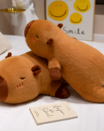 Two brown chonky sleeping capybara plushies resting on a white bed with a book. Perfect gift!