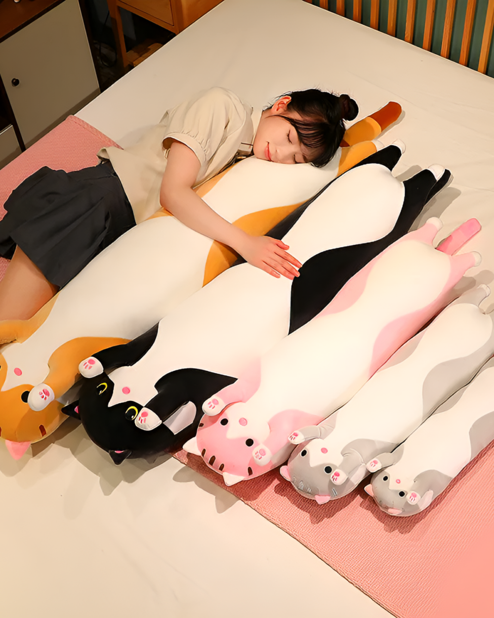A person sleeps with a Cloud Kitty Long Snuggle Pillow, a cute long cat plush. Several other cat plushes are nearby.