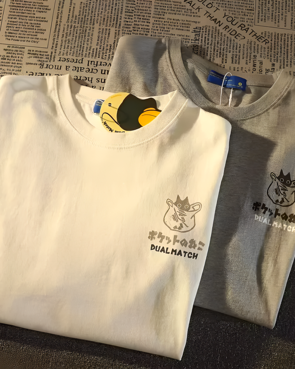 A white t-shirt featuring a Dual Match Neko Bag graphic design. The design includes cartoon cats and text on a patterned background.
