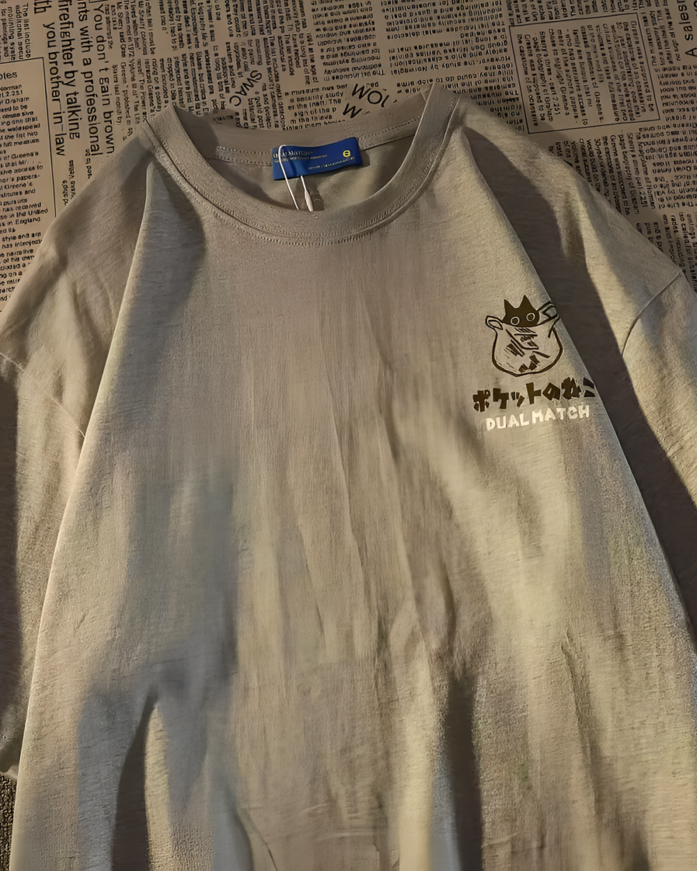 Close-up of a black Dual Match Neko Bag T-Shirt placed on newspapers. The shirt features a graphic design.