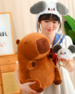 A woman holding a brown capybara plushie and a smaller squeaky capybara plushie wearing a hat. They’re the Hatted Capybara Plushies - Squeaky Purse Edition.