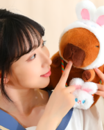Woman interacting with a brown capybara plushie wearing a hat and with paw details. Part of the Hatted Capybara Plushies - Squeaky Purse Edition.