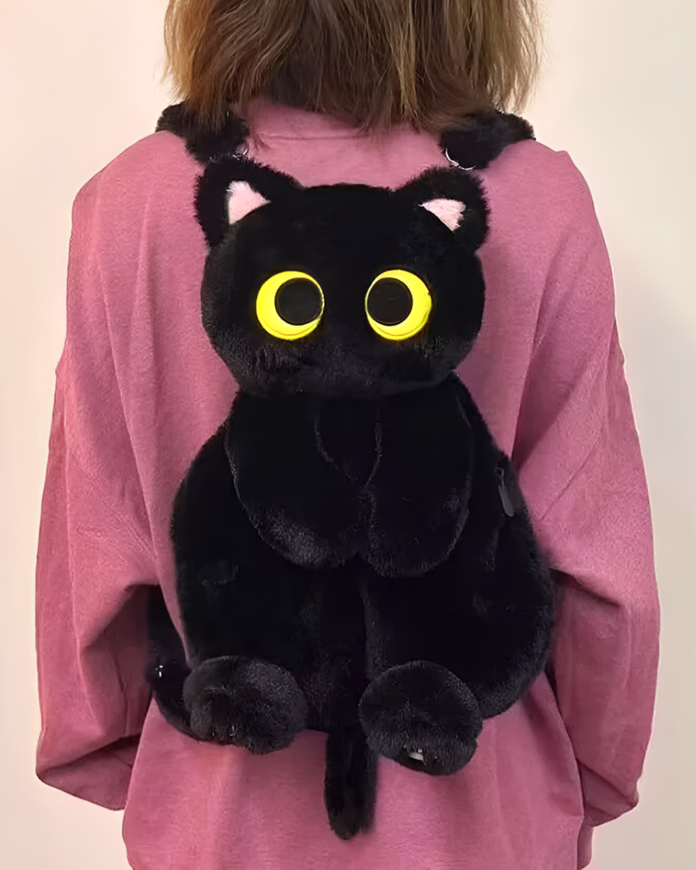 Person wearing a pink sweatshirt with a black Kawaii Cat Plushie Backpack attached. The backpack features large yellow eyes.