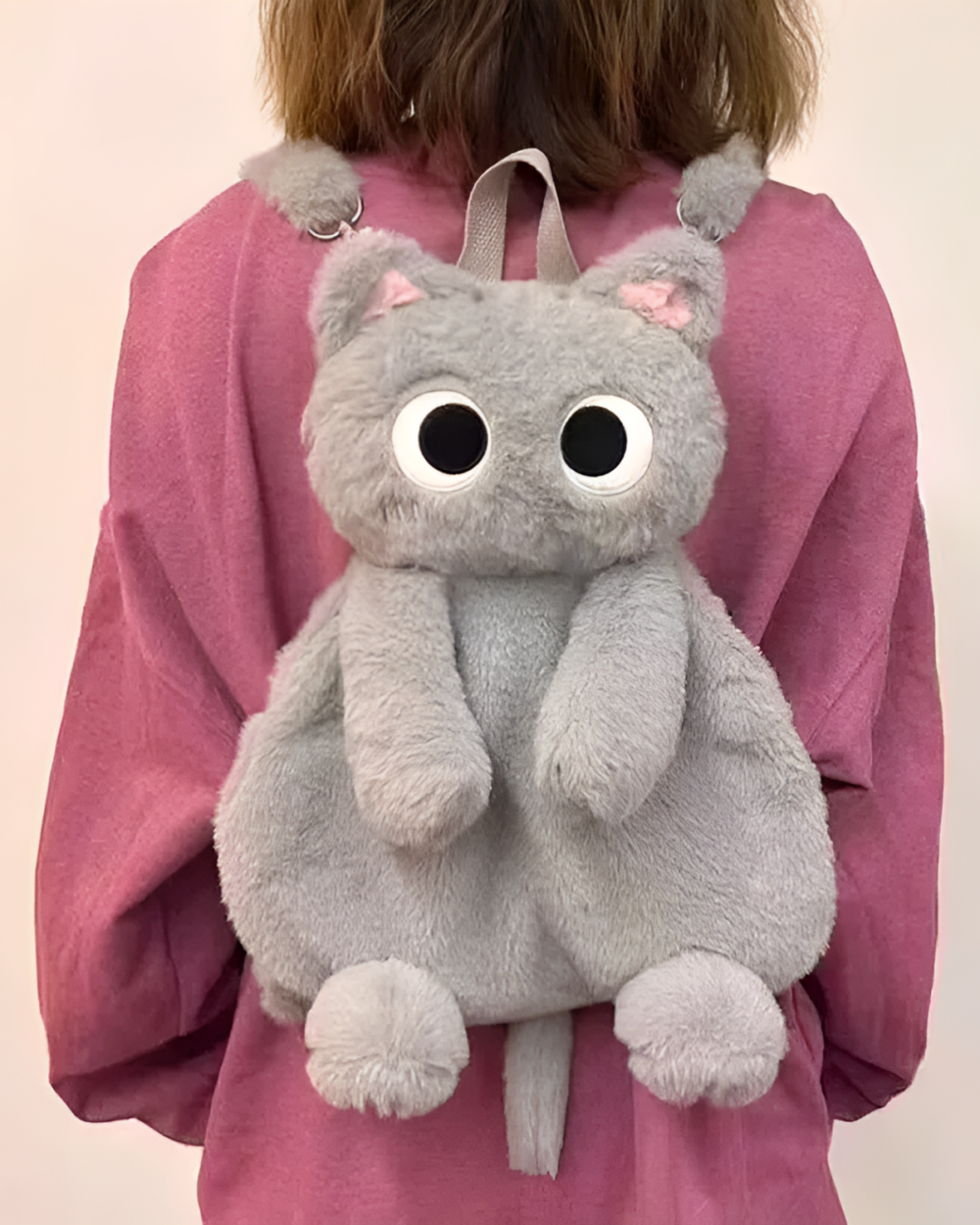 A person wearing a grey Kawaii Cat Plushie Backpack. The plushie features large eyes and a tail.