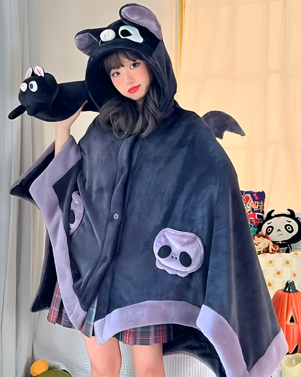 A woman wearing a black Kawaii Winged Eared Plush Cape and holding a matching plush bat. The cape features bat wings and a skull design.
