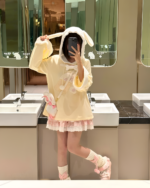A person wearing a beige jojifuku hoodie and matching socks in a restroom mirror reflection.
