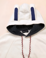 Close-up of a white bunny fleece cape with blue inner ears and a patterned hood lining. Includes braided drawstring ties.