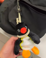 Hand holding a black and white plush Noosed Penguin Keychain with a red beak and yellow feet, attached to a bag.
