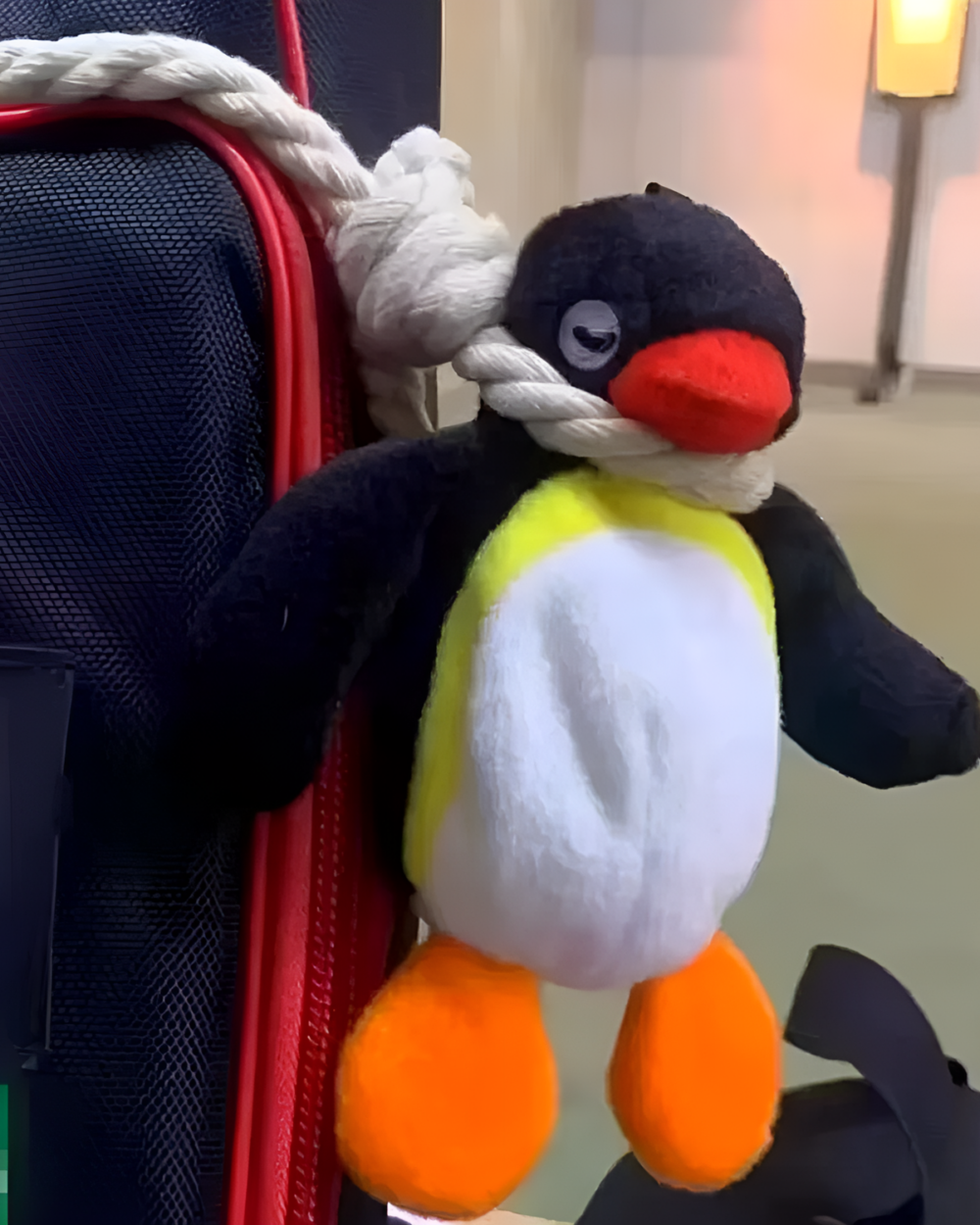 Close-up of a Noosed Penguin Keychain attached to a red backpack. The plush toy features black, white, and orange detailing.