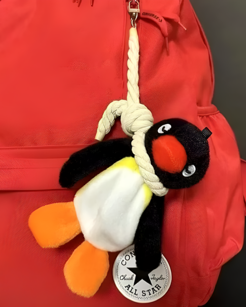 Close-up of a red backpack with a Noosed Penguin Keychain attached. The keychain features a black and white penguin plush.