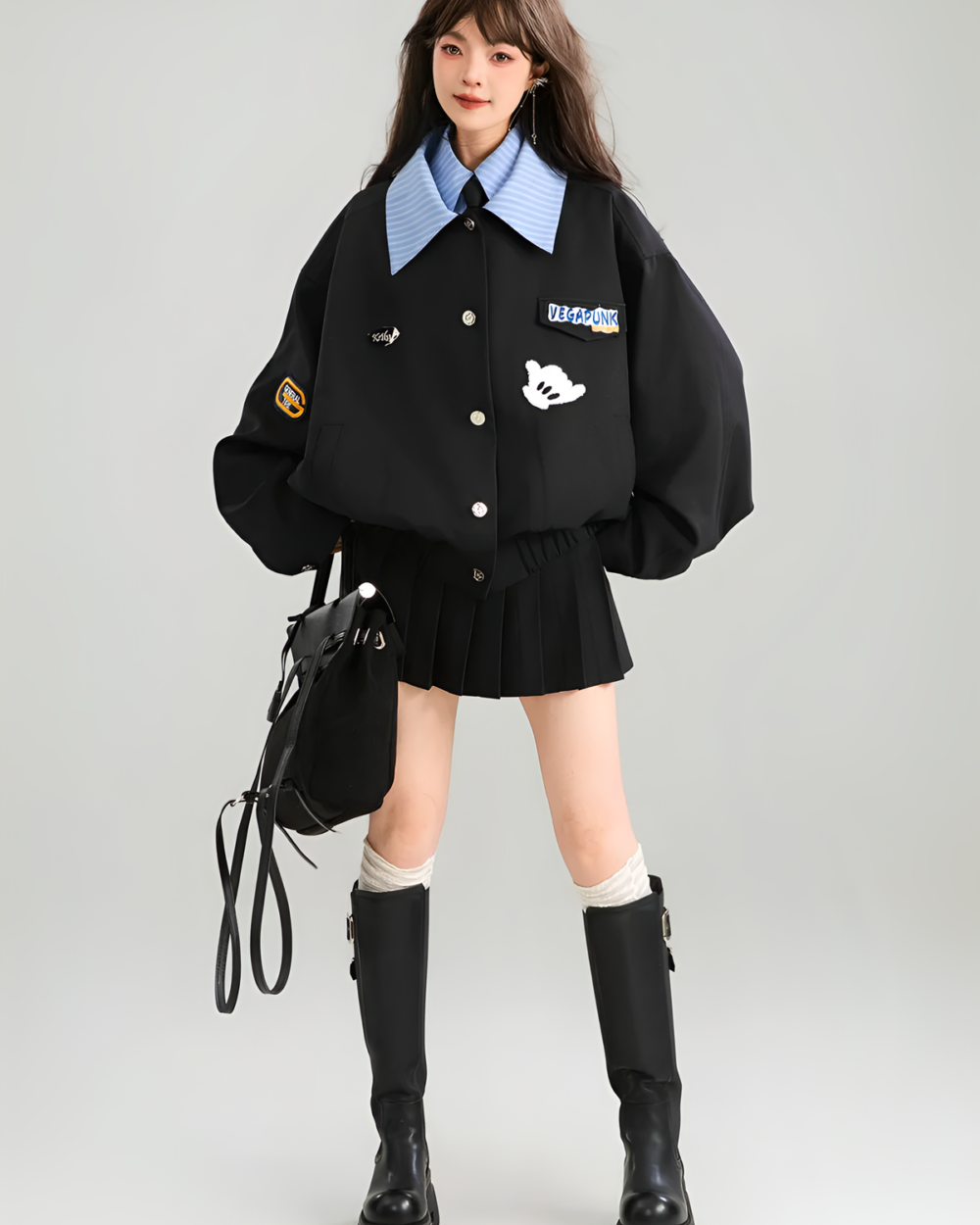 Woman wearing a black Patchy Jacket with decorative patches and a blue shirt underneath, paired with a pleated skirt and boots.