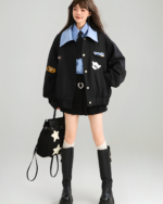 Woman wearing a black Patchy Jacket with embroidered patches and button closures, styled with a blue shirt and black boots.