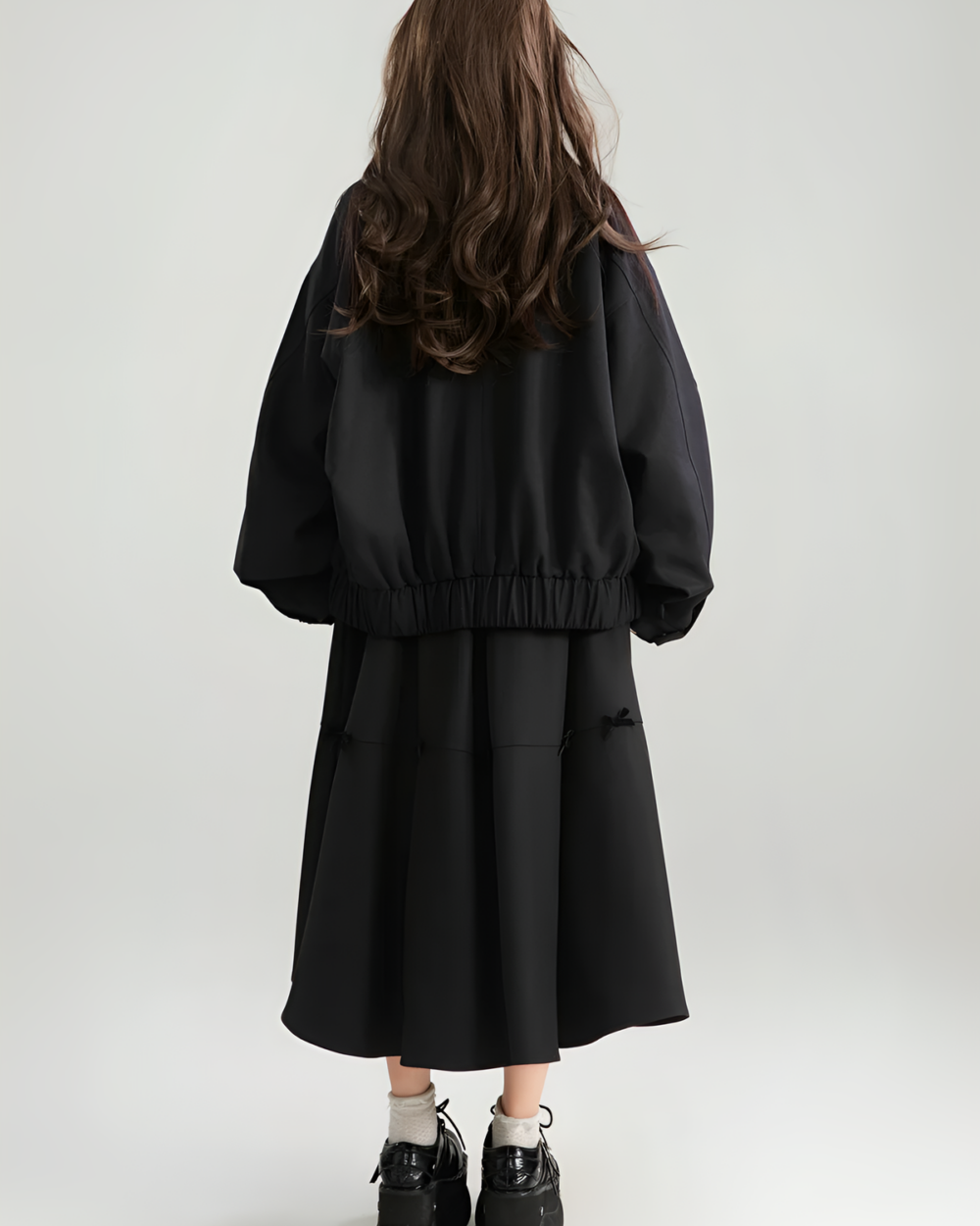 Rear view of a woman wearing a black Patchy Jacket with a preppy aesthetic. The jacket has a long line and elasticated cuffs.