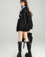 Woman wearing a black Patchy Jacket with preppy aesthetic, and black boots. The jacket features colorful patches.