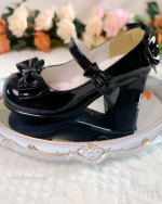Black Petal Block-Heel Mary Janes with a bow detail, displayed on a decorative plate. Shiny patent leather finish.