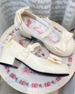 Close-up of white Petal Block-Heel Mary Janes with a bow detail and a block heel. The shoes are on a patterned surface.
