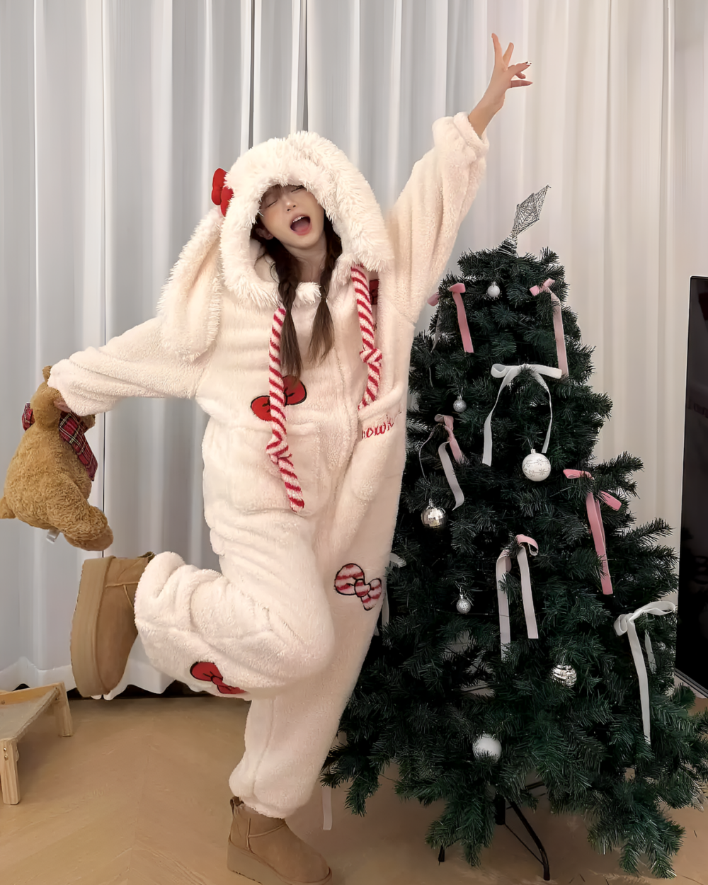 Woman wearing a cozy pink Slumber Bow-Bunny Ribbon bunny kigurumi pajamas near a decorated Christmas tree.