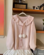 Pink soft girl sweater hangs on a hanger, featuring a fluffy collar and pom-poms.