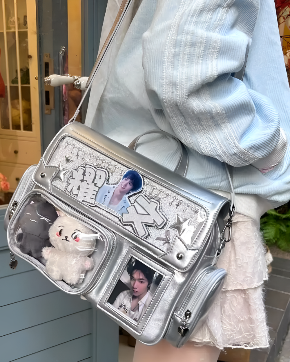 Silver ita bag with clear pockets displaying photos and plushies, worn over a light blue sweater. Korean Edition.