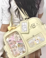 A person wearing a beige Strapped JK Ita Bag with a clear front panel displaying collectible cards and keychains.