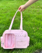 Pink Strapped JK Ita Bag with transparent window, buckles, and adjustable straps shown outdoors on grass.
