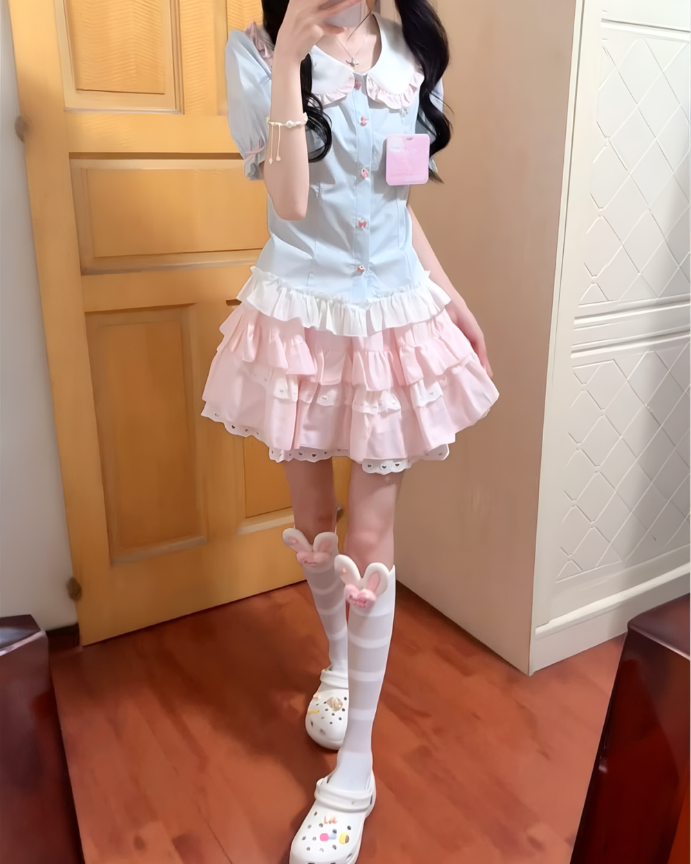 A person wearing a Sweet Lolita School Suit, featuring a light blue dress with layered ruffles and pink accents.