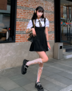 Woman modeling a black suspender skirt set with a white blouse and platform boots.