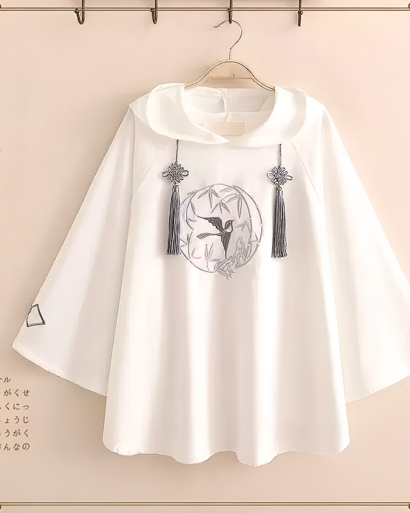 White hooded poncho with embroidered hummingbird design, Zephyr Bamboo Hooded Poncho. A detailed image of the garment hanging on a rack.