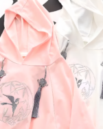 Close-up of Zephyr Bamboo hooded poncho in pink and white with embroidered bird details and tassel ties.